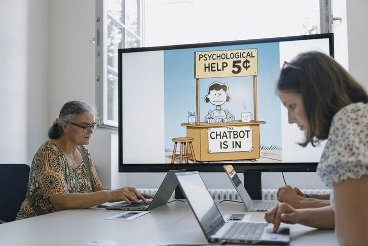 Zwei Frauen arbeiten an Laptops. Im Hintergrund zeigt ein Bilschirm eine Folie zum Thema Chatbot. 