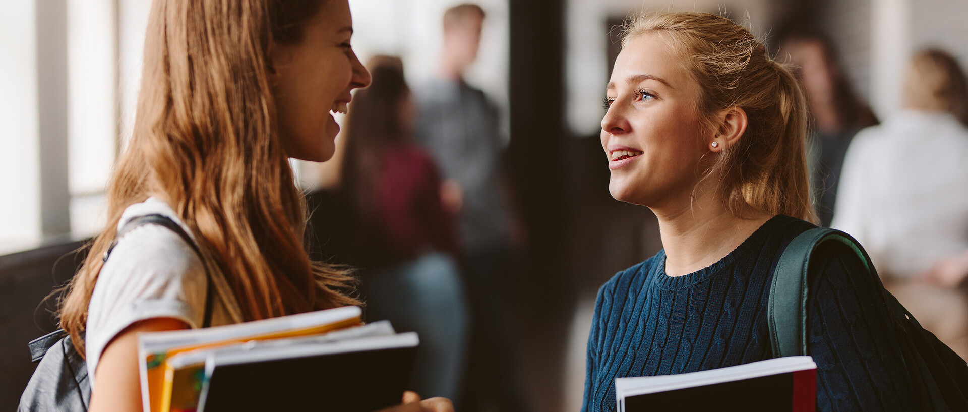 Zwei Studentinnen unterhalten sich stehend miteinander und halten Unterlagen in der Hand.