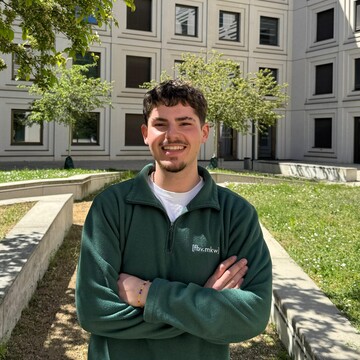 Lex has short brown hair and is wearing a green sweatshirt. His arms are crossed and he is smiling at the camera.