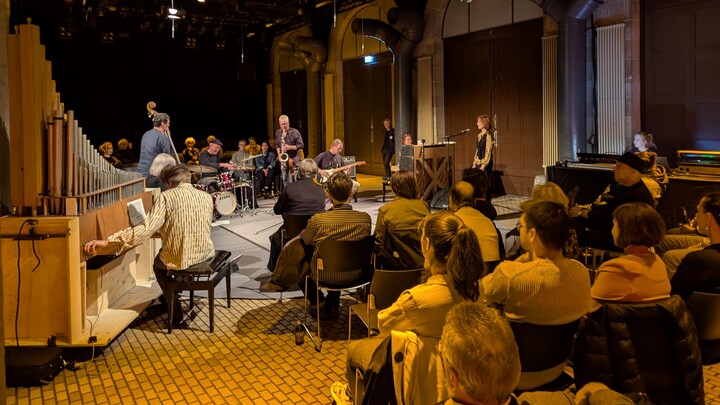 Eindruck des Konzerts. Im rechten Vordergrund ist das Publikum zu sehen, das zu den Künstler:innen in der Mitte der Konzerthalle blickt.