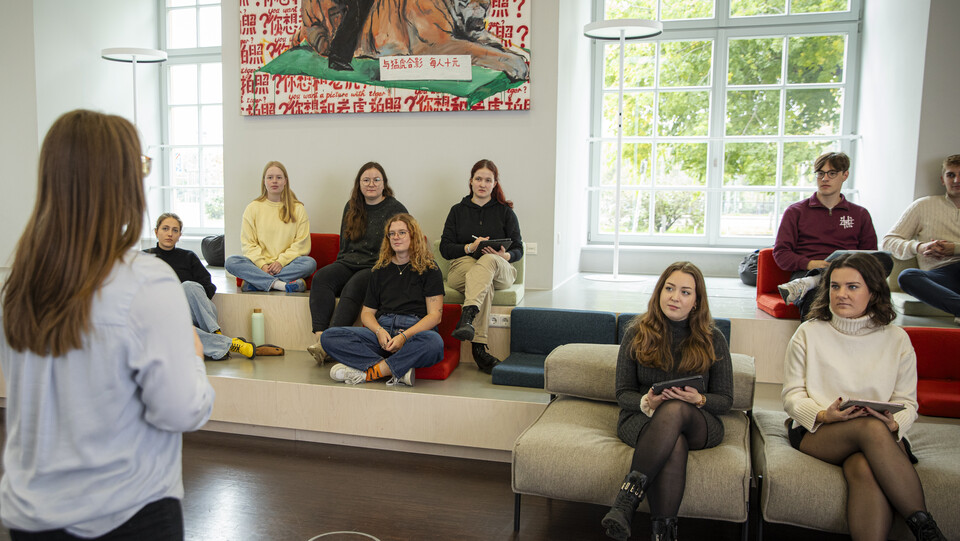 Eine Frau steht mit dem Rücken zur Kamera und spricht zu einer Gruppe von Studierenden, die in einem hellen, modernen Seminarraum auf gestuften Sitzflächen und Sesseln sitzen. Einige halten Tablets oder Notizblöcke in der Hand und hören aufmerksam zu.