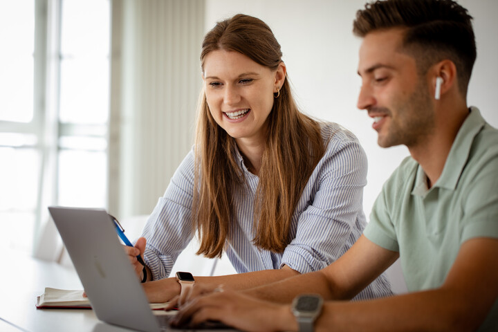 Eine junge Frau und ein junger Mann sitzen lachend vor einem Laptop.