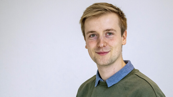 Jan Kemper. He is wearing a green sweater over a blue shirt and is standing in front of a greyish wall.