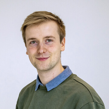 Jan Kemper. Er trägt einen grünen Pullover über einem blauen Hemd und steht vor einer weißen Wand.