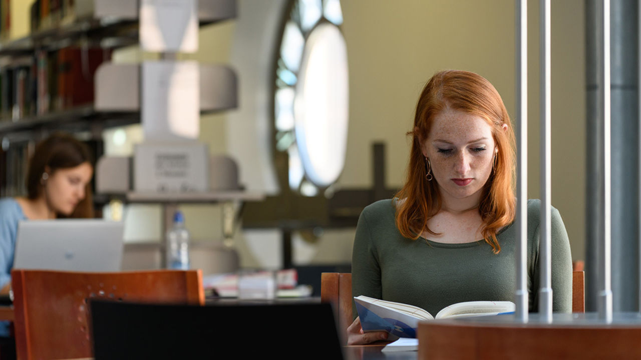Eine Studentin sitzt in der Bibliothek und liest ein Buch. Sie hat kupferrotes Haar und trägt ein dunkelgrünes Oberteil.