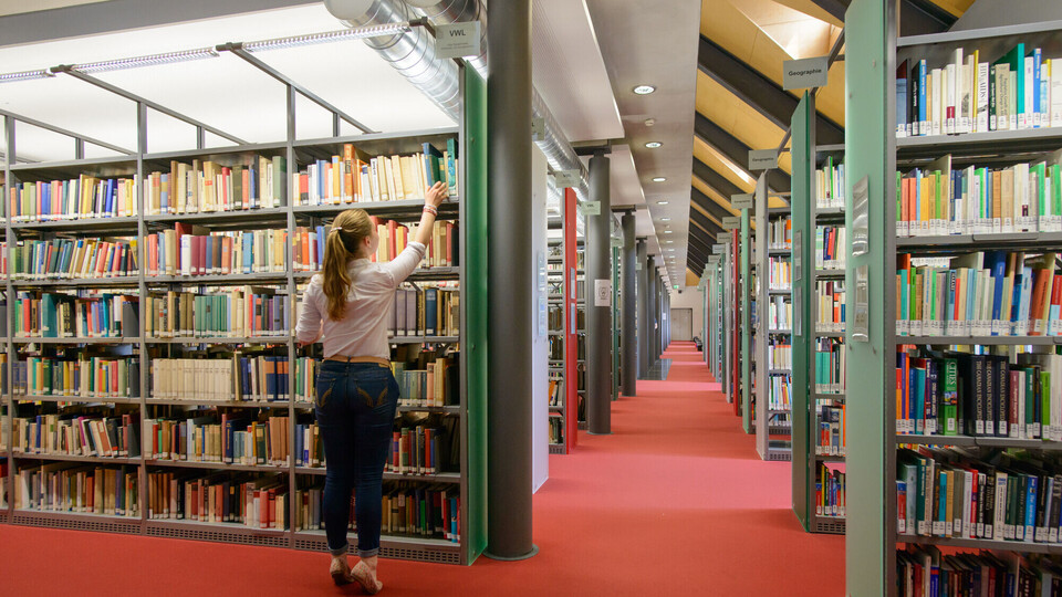 Eine Studentin stellt sich auf die Zehenspitzen, um aus dem obersten Bibliotheksregal ein Buch zu ziehen.