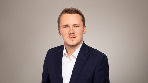 Andreas Hamann. He is wearing a black suit and is standing in front of a grey photowall.