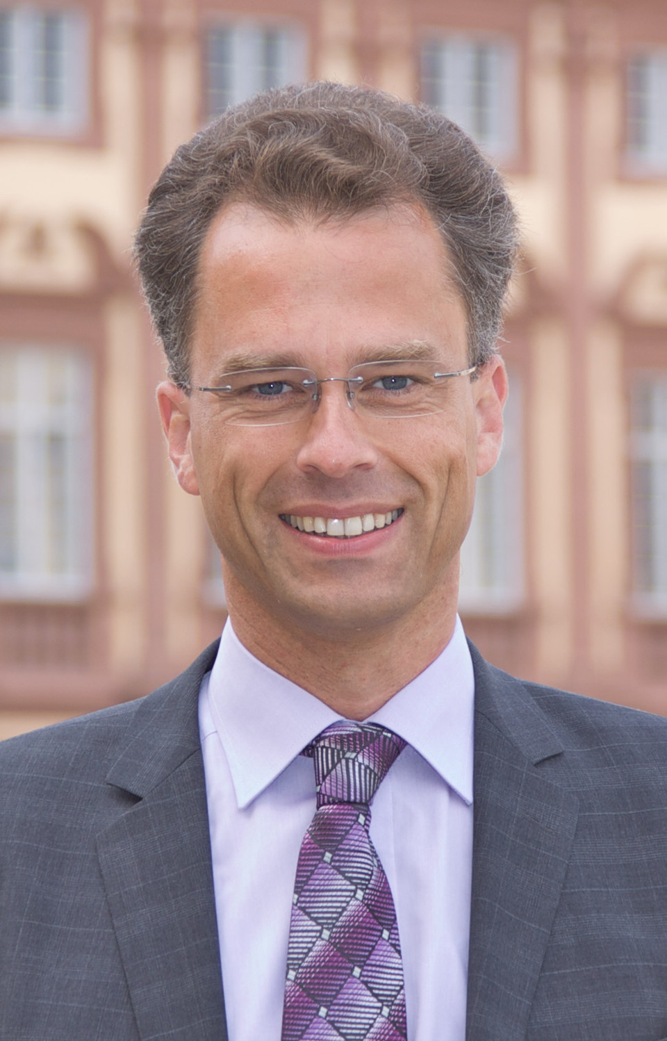 Portrait von Georg Bitter. Er tträgt ein weißes Hemd, eine lila-farbene Krawatte sowie einen grauen Blazer und steht vor dem Schloss.