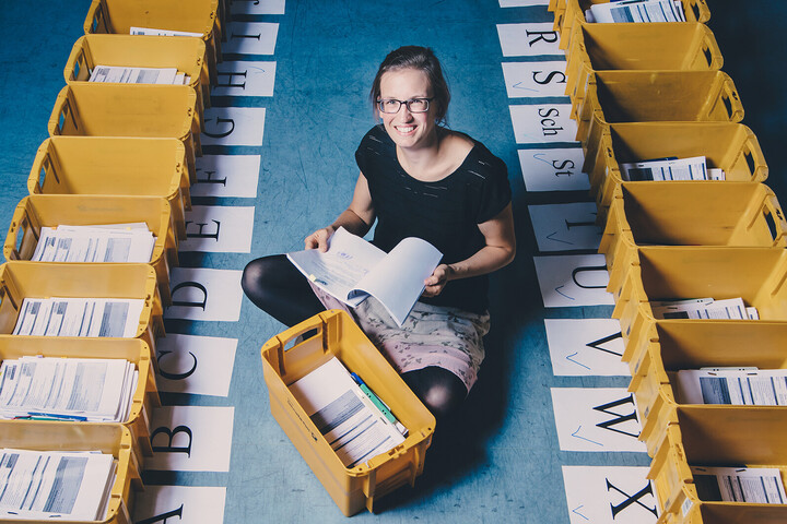 Eine Mitarbeitende der Universität Mannheim sitzt auf dem Boden. Um sie herum stehen gelbe Postkisten. Die Kisten sind mit verschiedenen Buchstaben beschriftet.