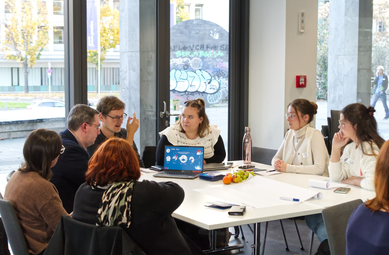 Ein Referent präsentiert den Teilnehmenden mit seinem Laptop sein Projekt. Gemeinsam sitzen alle um einen Tisch herum.