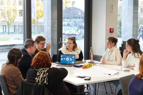 Ein Referent präsentiert den Teilnehmenden mit seinem Laptop sein Projekt. Gemeinsam sitzen alle um einen Tisch herum.
