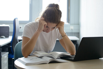 Studentin sitzt verzweifelt über einem Buch am Schreibtisch mit Laptop.