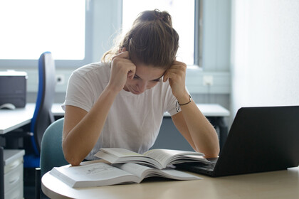Studentin sitzt verzweifelt über einem Buch am Schreibtisch mit Laptop.