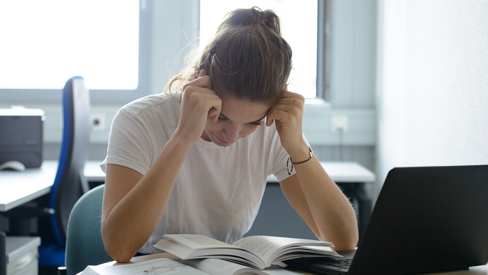 Studentin sitzt verzweifelt über einem Buch am Schreibtisch mit Laptop.