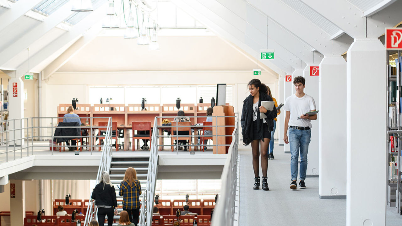 Studierende arbeiten im Bibliotheksbereich A3. Die Tische und Stühle sind dunkelrot. Durch das Glasdach dringt viel Licht in das Gebäude.