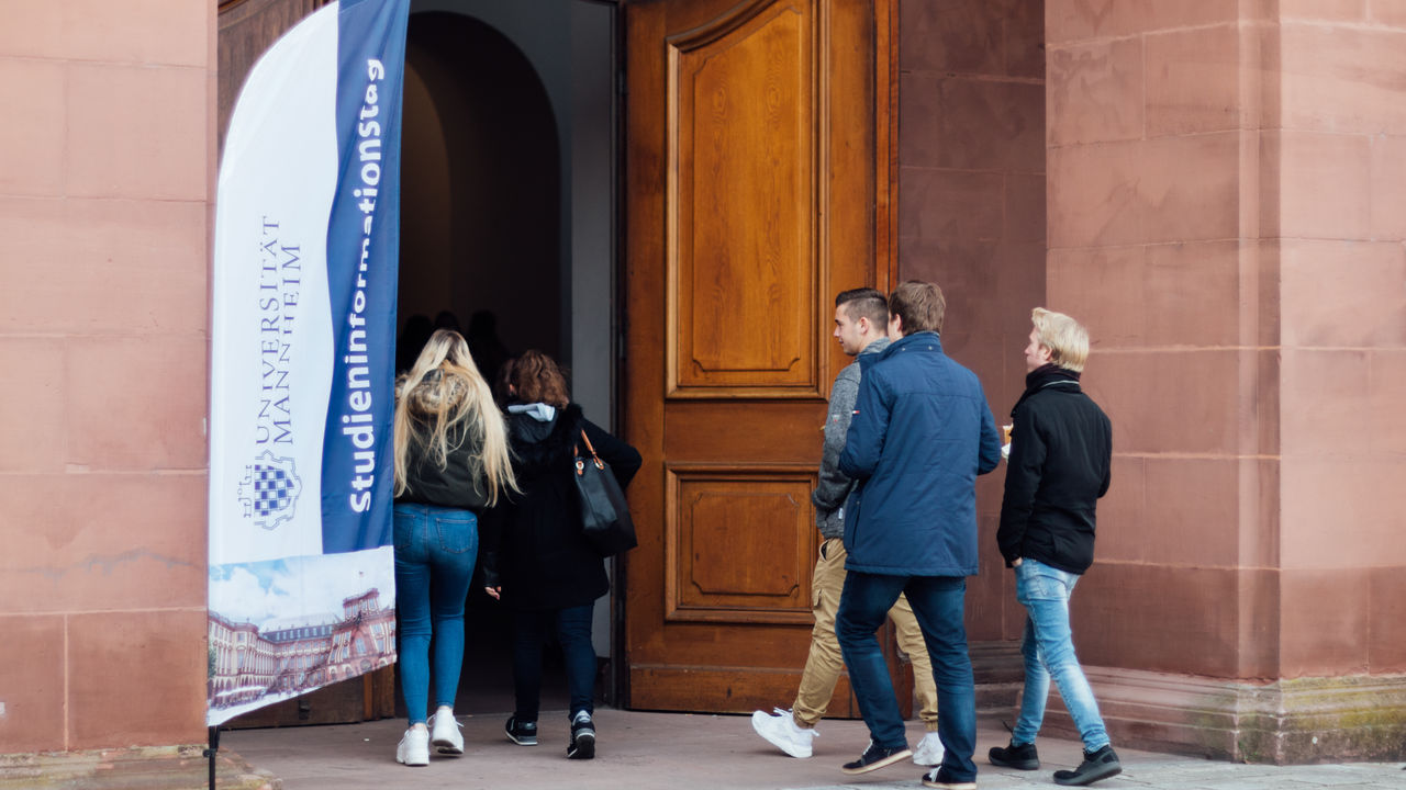 Studierenden gehen durch eine große Holzpforte in die Uni. Neben der Tür ein Banner mit der Aufschrift "Studieninformationstag".