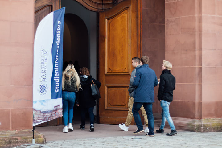Studierenden gehen durch eine große Holzpforte in die Uni. Neben der Tür ein Banner mit der Aufschrift "Studieninformationstag".