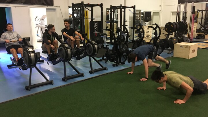 Drei Männer trainieren im Fitnessstudio an Rudergeräten, daneben machen zwei Männer Körpergewichtsübungen