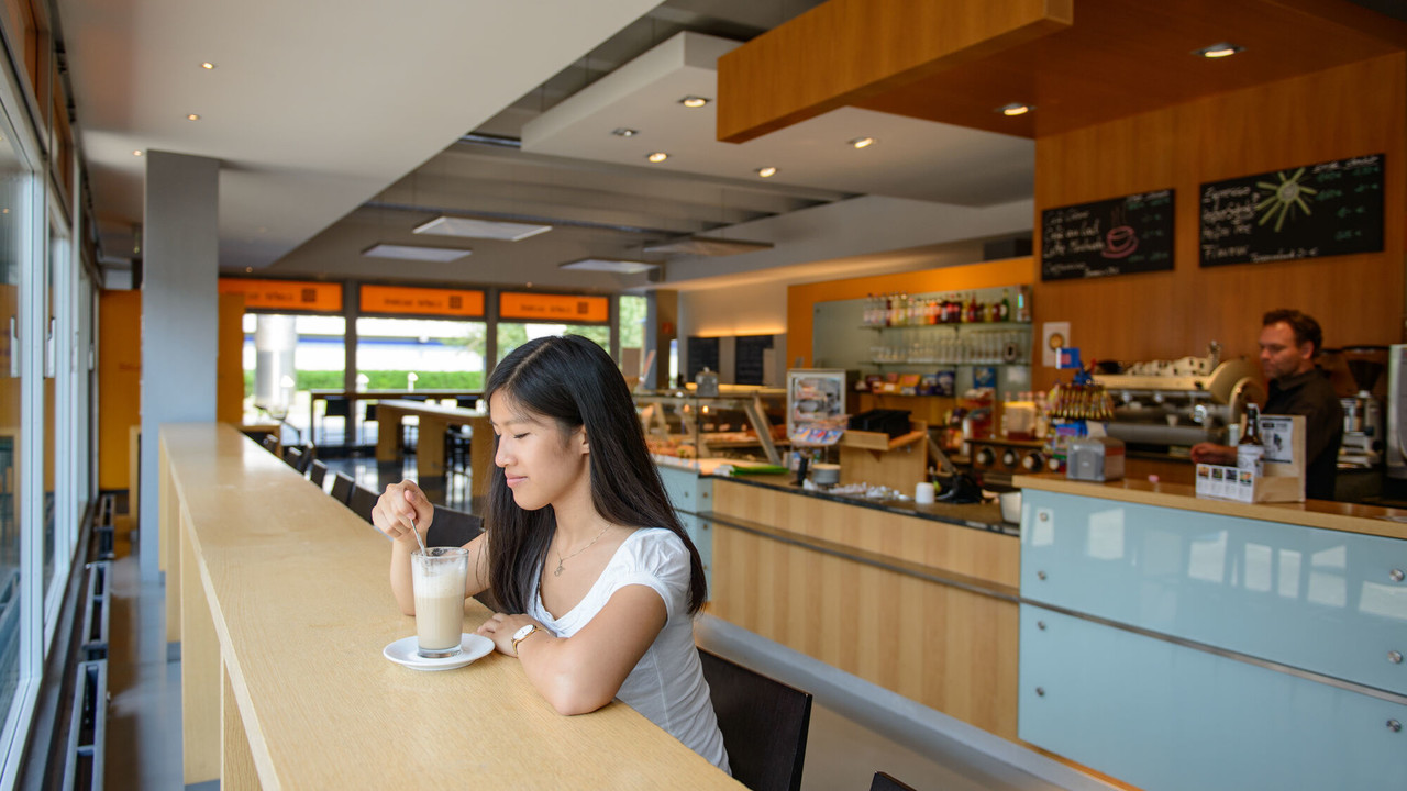 Eine Studentin trinkt einen Cappuccino im "Café Soleil" der Universität Mannheim und im Hintergrund steht ein Mitarbeiter hinter dem Thresen.
