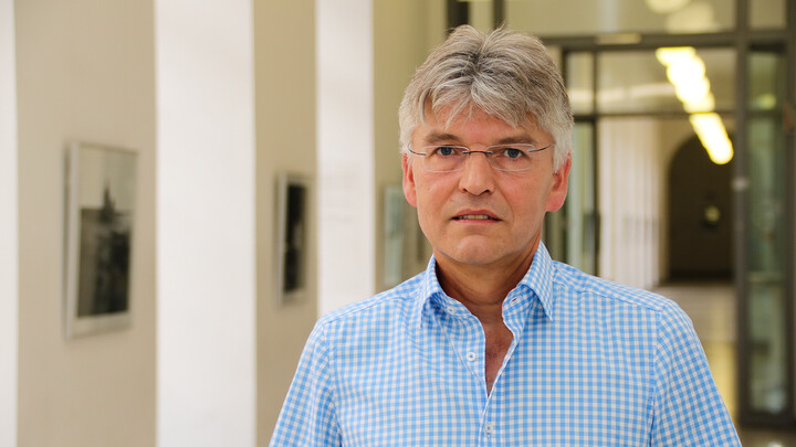 Portraitfoto eines Mannes mit blau-weiß kariertem Hemd. Im Hintergrund ist ein langer Gang mit mehreren Bildern