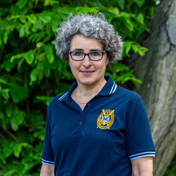Susanne Quincke steht mit blauem Polo-Shirt vor einer grünen Blätterwand und lächelt in die Kamera.