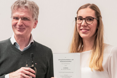 Lea Backes hält die Auszeichnung für ihre wissenschaftliche Arbeit lächelnd hoch. Neben ihr steht lächeln Professor Justus Fetscher.