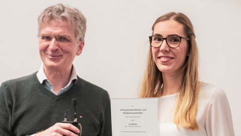 Lea Backes hält die Auszeichnung für ihre wissenschaftliche Arbeit lächelnd hoch. Neben ihr steht lächeln Professor Justus Fetscher.