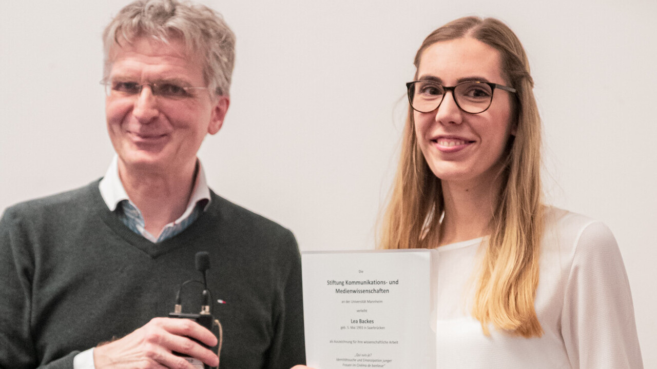Lea Backes hält die Auszeichnung für ihre wissenschaftliche Arbeit lächelnd hoch. Neben ihr steht lächeln Professor Justus Fetscher.
