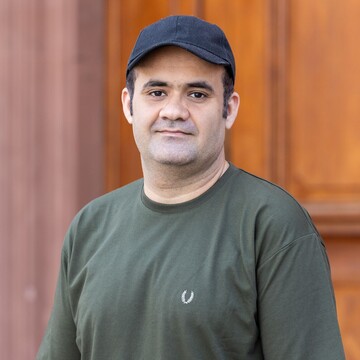 Zubair hat kurze Haare. Die Person trägt eine Schildmütze und ein olivgrünes Shirt. Er steht vor einer Holztür und schau in die Kamera.
