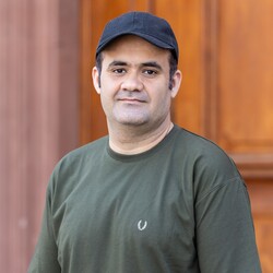 Zubair hat kurze Haare. Die Person trägt eine Schildmütze und ein olivgrünes Shirt. Er steht vor einer Holztür und schau in die Kamera.