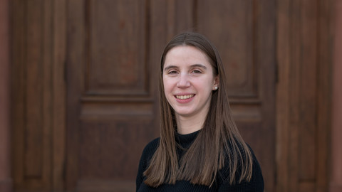 Jasmina Simon. She is wearing a black sweater and is standing in front of a brown door.