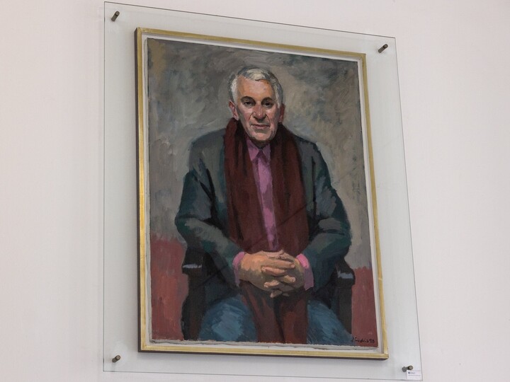 Porträt von Herbert Prechel: Er trägt ein rosa Hemd, ein grauem Jackett und einem langen roten Schal. Er sitzt auf einem Stuhl mit ineinander verschränkten Händen.