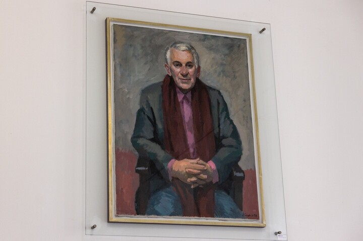 Porträt von Herbert Prechel: Er trägt ein rosa Hemd, ein grauem Jackett und einem langen roten Schal. Er sitzt auf einem Stuhl mit ineinander verschränkten Händen.