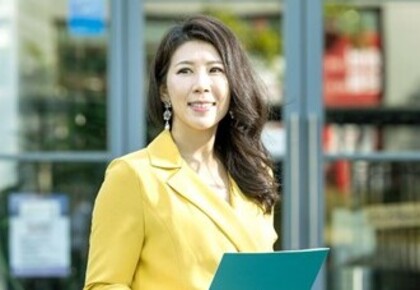 Meeyoung Cha im Freien vor einer Glastür stehend. Sie trägt einen gelben Blazer und hält ein grünes Klemmbrett in den Händen.