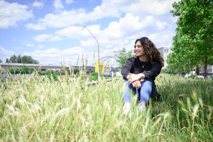 Eine Frau sitzt lächelnd im hohen Gras vor Stadtlandschaft mit Brücke und Gebäuden im Hintergrund.