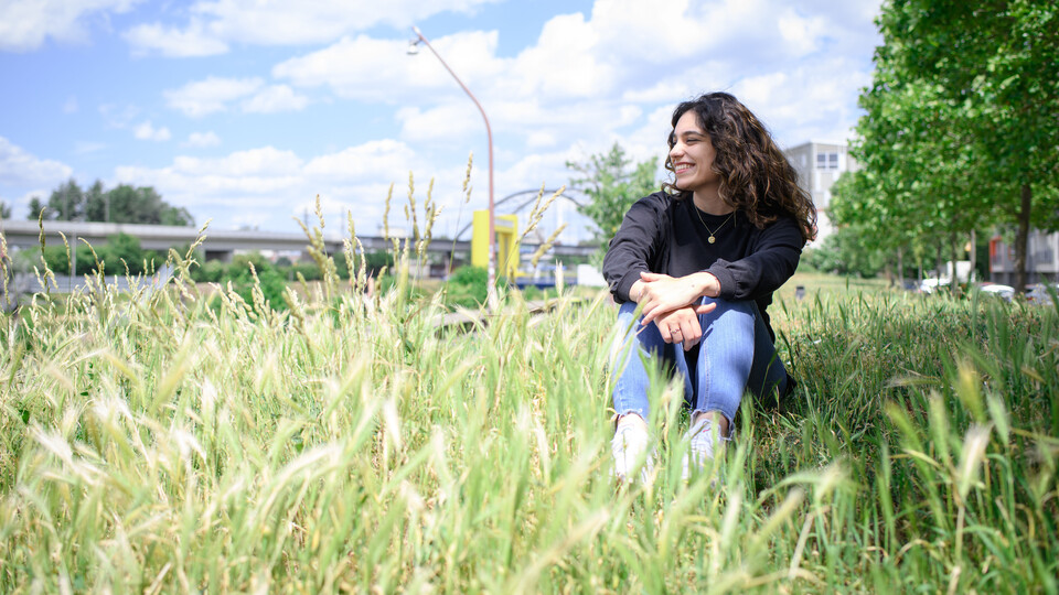 Eine Frau sitzt lächelnd im hohen Gras vor Stadtlandschaft mit Brücke und Gebäuden im Hintergrund.