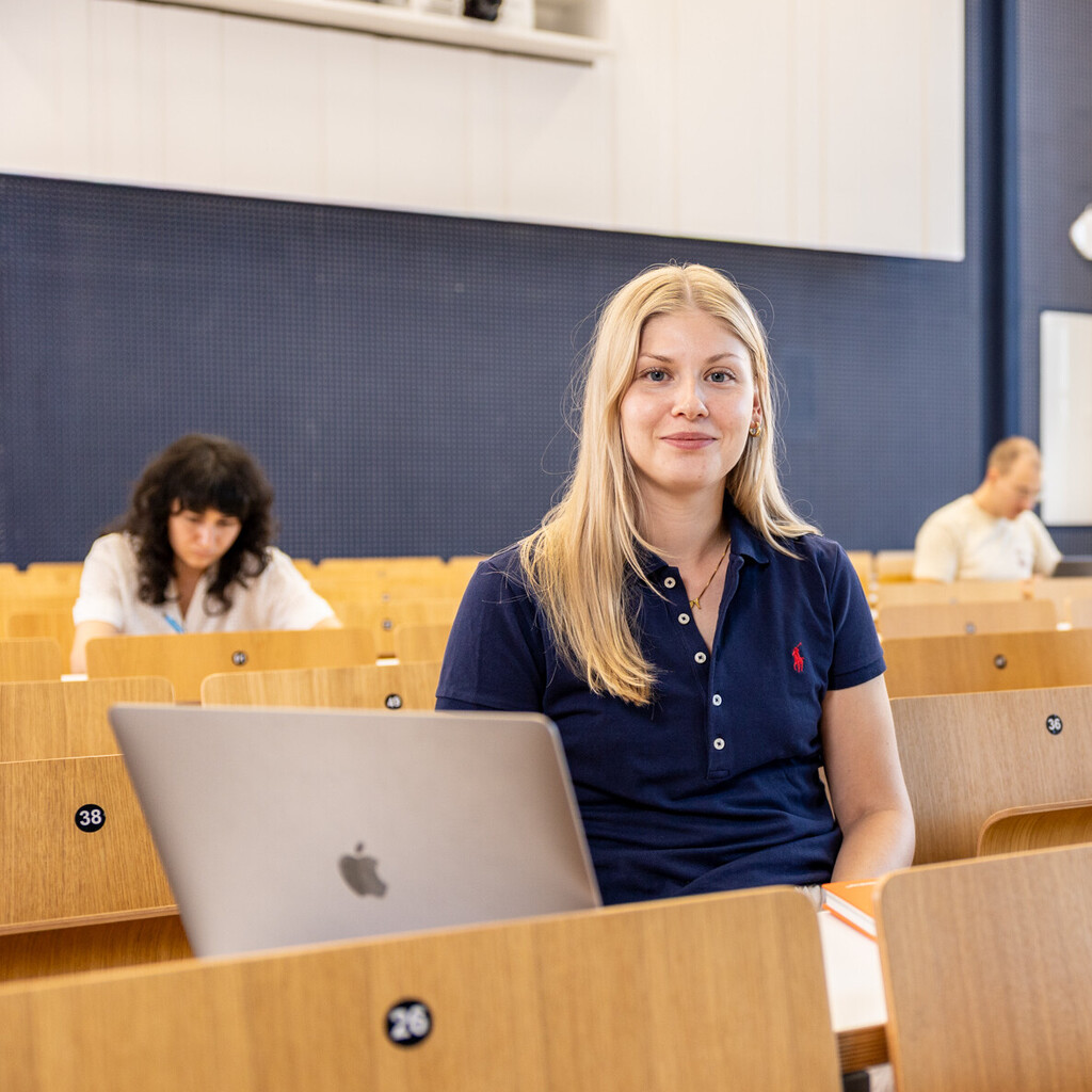 Eine Studentin sitzt in einer Bankreihe im Hörsaal. Vor sich hat sie ihren aufgeklappten Laptop. Sie schaut direkt in die Kamera und lächelt. Im Hintergrund sitzen weitere Studierende.
