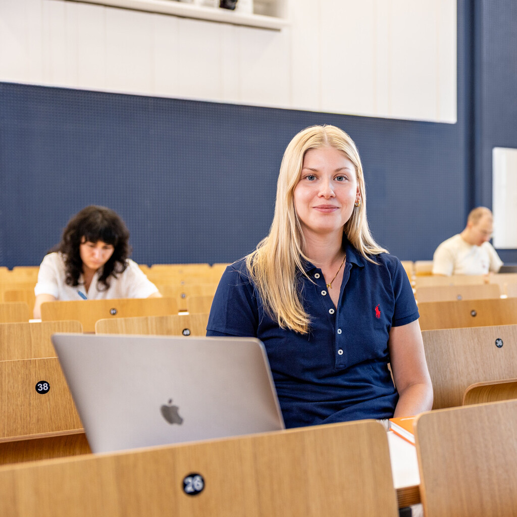 Eine Studentin sitzt in einer Bankreihe im Hörsaal. Vor sich hat sie ihren aufgeklappten Laptop. Sie schaut direkt in die Kamera und lächelt. Im Hintergrund sitzen weitere Studierende.