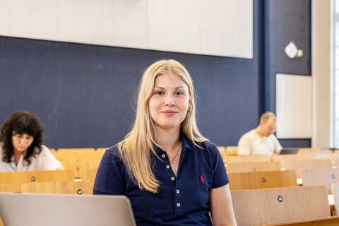 Eine Studentin sitzt in einer Bankreihe im Hörsaal. Vor sich hat sie ihren aufgeklappten Laptop. Sie schaut direkt in die Kamera und lächelt. Im Hintergrund sitzen weitere Studierende.