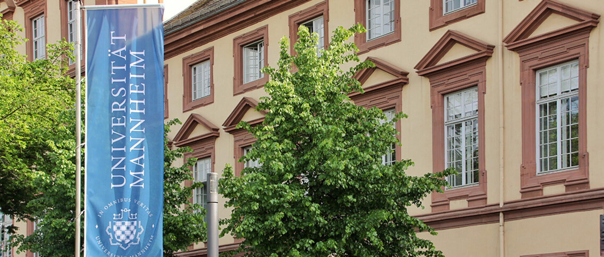 Eine blaue Fahne mit dem Logo der Uni Mannheim weht vor dem Haupteingang der Universität im Ostflügel des Schlosses.