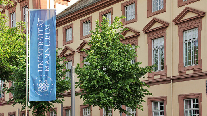 Eine blaue Fahne mit dem Logo der Uni Mannheim weht vor dem Haupteingang der Universität im Ostflügel des Schlosses.