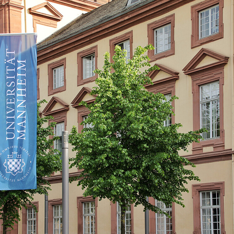 Eine blaue Fahne mit dem Logo der Uni Mannheim weht vor dem Haupteingang der Universität im Ostflügel des Schlosses.