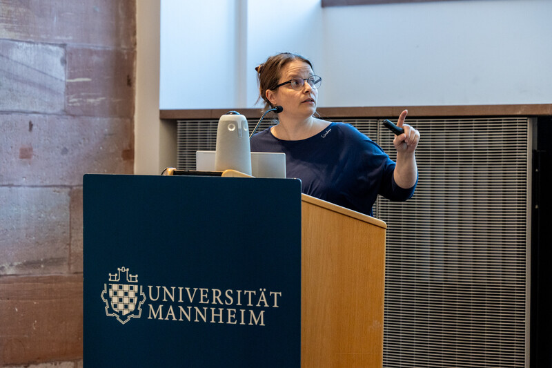 Dr. Jessica Steinberger steht am Pult der Bühne während der Hauptveranstaltung der Diversity Week 2025. Sie gestikuliert in Richtung der Powerpoint Folie, die jedoch nicht zu sehen ist. Sie hat die Haare hochgesteckt, trägt ein dunkelblaues T-Shirt und eine Brille.