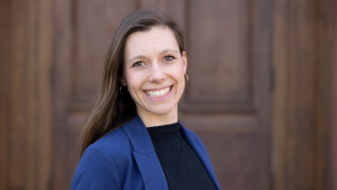 Marina Hoch. She is wearing a dark blue jacket over a black sweater and is standing in front of a brown door.