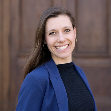 Marina Hoch. Sie trägt ein königsblaue Jacke über einem schwarzen Pullover und steht vor einer Holztür.