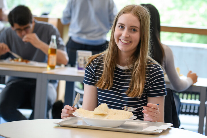 Eine Studentin sitzt mit Messer und Gabel in der Hand an einem Tisch in der Mensa und lächelt, auf ihrem Teller liegt ein Wrap.