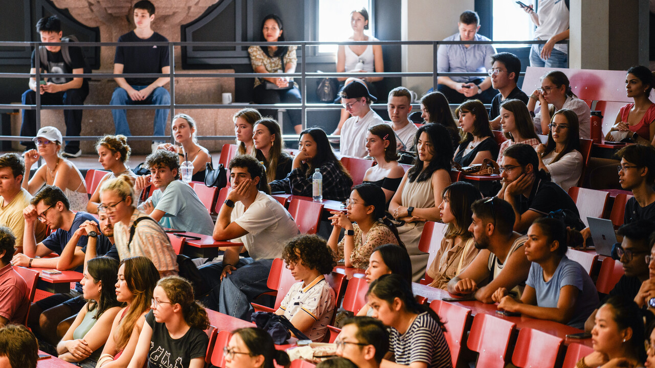 Viele Studierende sitzen in einem Hörsaal mit roten Möbeln.