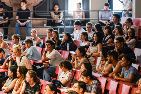 Viele Studierende sitzen in einem Hörsaal mit roten Möbeln.