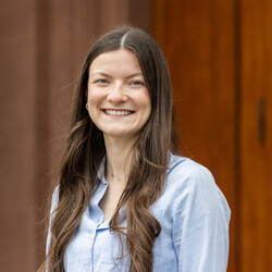 Porträt von Evelyn Bentz. Evelyn Bentz trägt eine hellblaue Bluse und lange braune, gelockte Haare. Ohrringe und Halskette sind dezent. Sie lacht offenen in die Kamera.