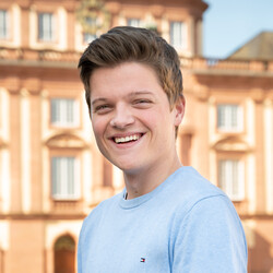 Georg Schade steht lachend vor dem Mannheimer Schloss. Er hat kurze braune Haare und trägt ein hellblaues Langarm-Shirt.